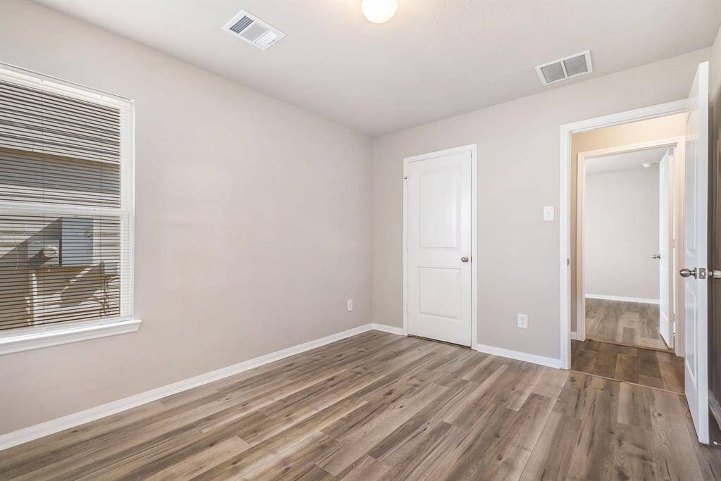 Another view of bedroom two, features neutral walls, wood-look flooring, and a window with blinds, creating a bright and versatile space. A door leads to an adjacent area, adding convenience. Ideal for a bedroom or home office.