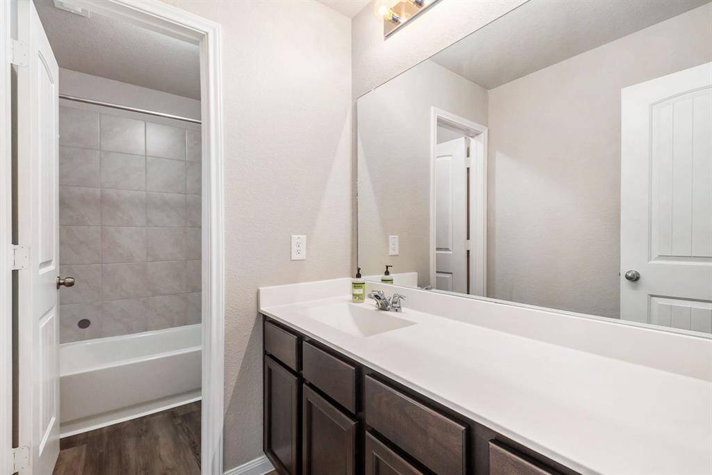 Shared bathroom features a sleek design with a long vanity, modern fixtures, and a large mirror. It includes a tub-shower combination with tiled walls, and the flooring is a stylish wood-look. Neutral tones create a clean, inviting space.