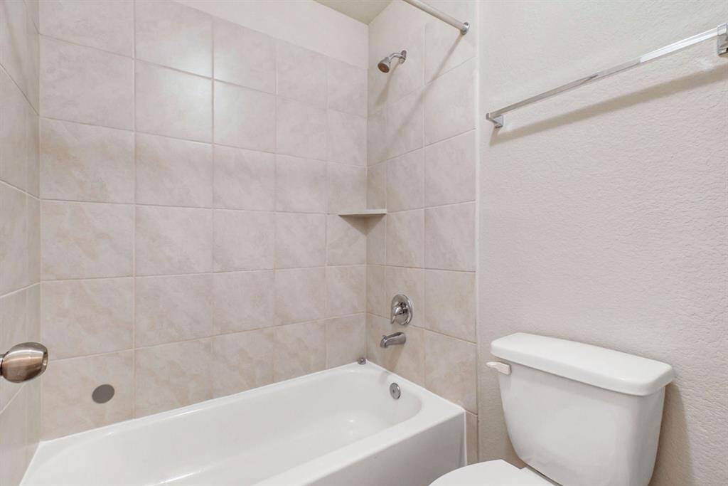 This bathroom features a clean, neutral-toned design with tiled walls surrounding a bathtub and shower combination. It includes a toilet and a towel bar, offering a simple, functional layout.