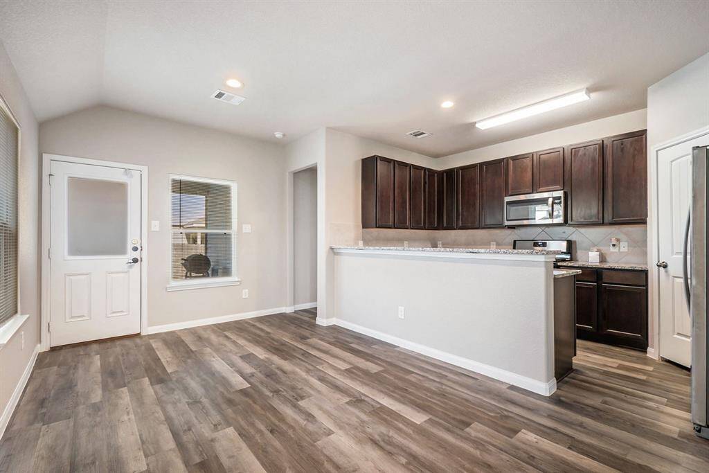 Modern kitchen with dark wood cabinets, granite countertops, and stainless steel appliances. The open layout connects to a living space with wood-style flooring and ample natural light from windows and a glass door.