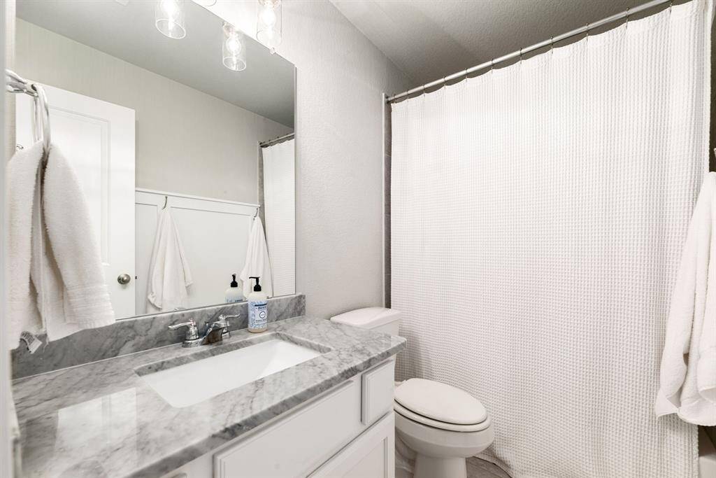 Secondary bath features tile flooring, bath/shower combo with tile surround, white cabinets, beautiful countertops, mirror, dark, sleek fixtures and modern finishes.