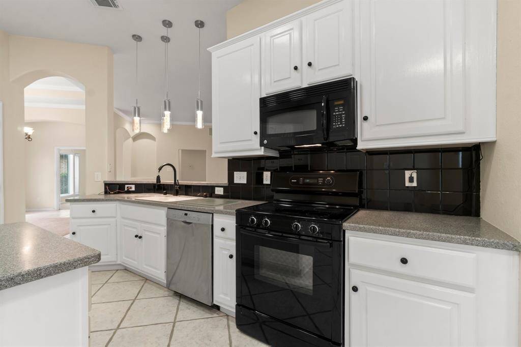 This spacious kitchen features modern black appliances, white cabinetry, and a central island. It's well-lit with sleek pendant lighting and has a neutral tile floor, creating a bright and inviting space.