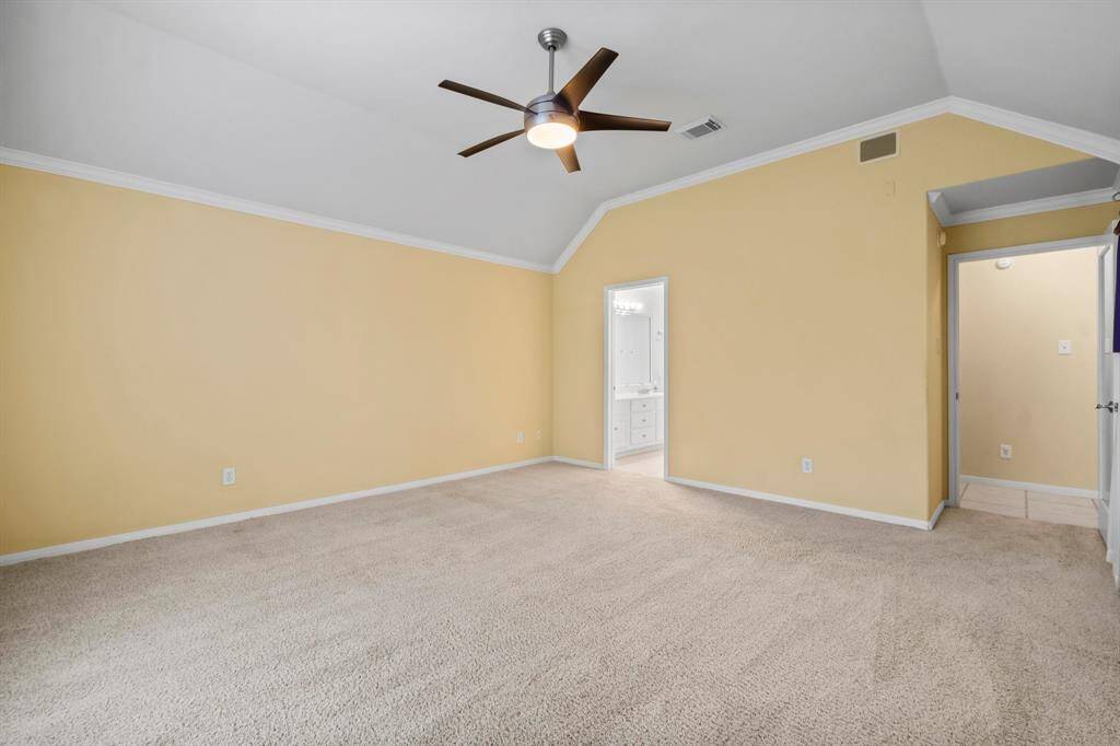 Spacious primary bedroom with vaulted ceiling, warm yellow walls, and plush carpeting. Features a large window with natural light and a modern ceiling fan.