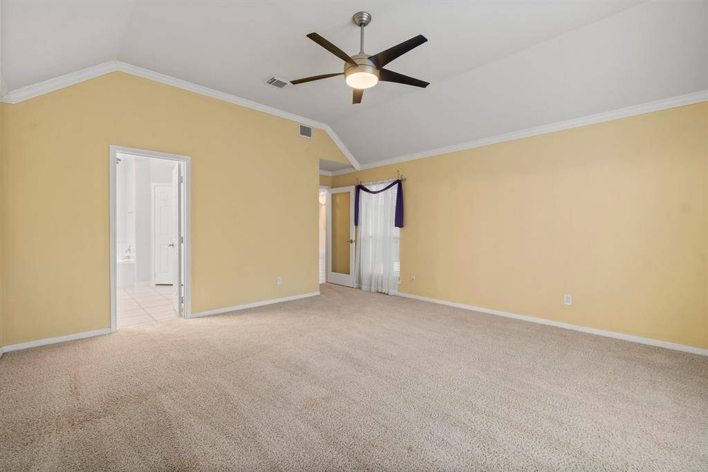 This spacious bedroom features soft yellow walls, plush carpeting, and a modern ceiling fan. It has an en-suite bathroom with a vanity, providing convenience and comfort.