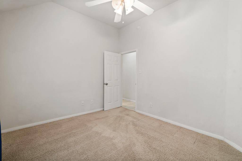 This room is bright and spacious with neutral walls and carpet, featuring a ceiling fan and a window with blue curtains. It includes a double-door closet, ideal for additional storage.