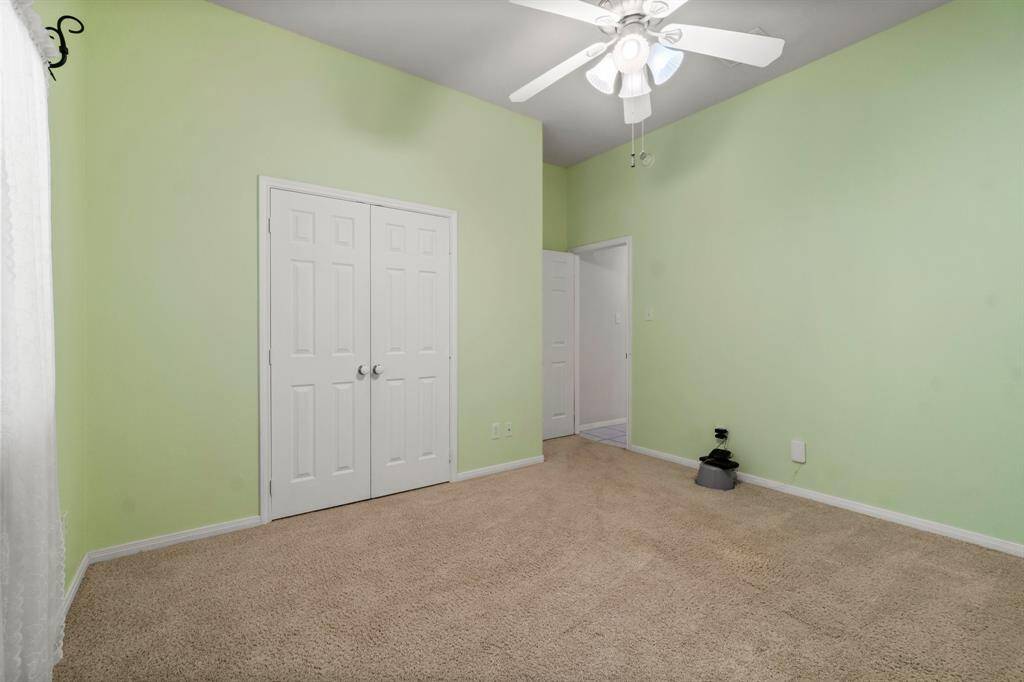 Bright, airy bedroom with soft green walls, carpeted floor, and a ceiling fan. Features a large window with sheer curtains, providing ample natural light.