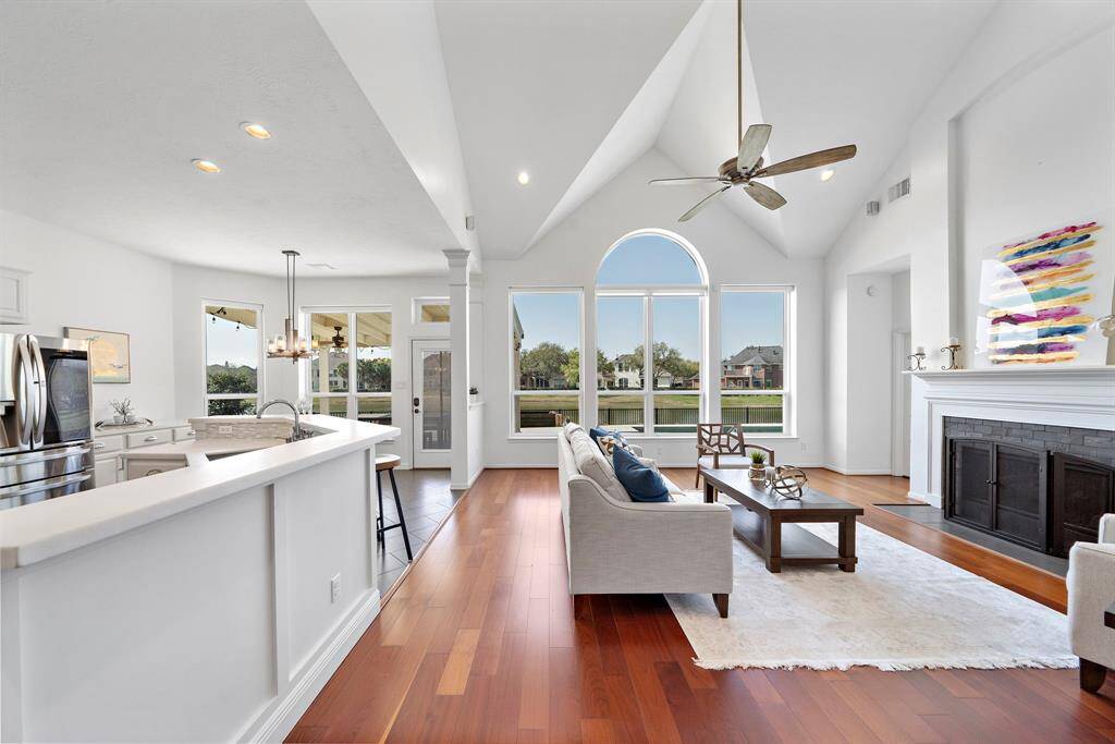 Bright and spacious open-concept living area with vaulted ceilings and large arched windows. The room features hardwood floors, a modern kitchen with stainless steel appliances, a cozy fireplace, and access to a patio with scenic views.