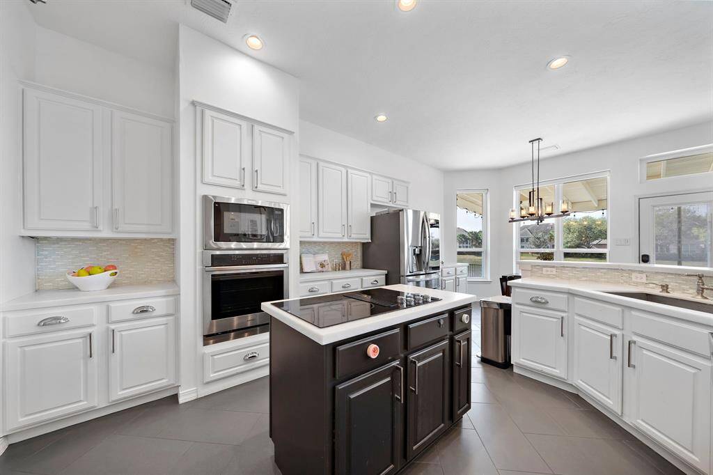 This modern kitchen features sleek white cabinetry, stainless steel appliances, and a central island with a cooktop. Large windows provide plenty of natural light, while a stylish chandelier adds elegance. Ideal for cooking and entertaining.