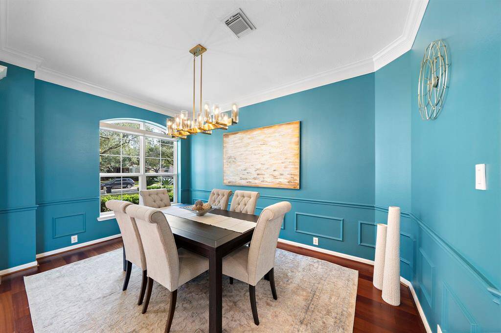This dining room features elegant blue walls, a modern chandelier, and a large window providing natural light.