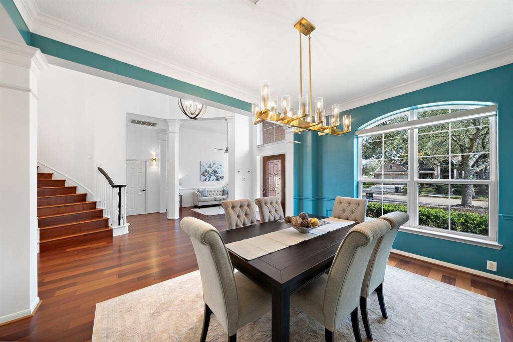 A spacious and elegant dining area with a large window offering plenty of natural light. The room features a stylish chandelier, hardwood floors, and a view of a cozy living space. A staircase leads to the upper level.