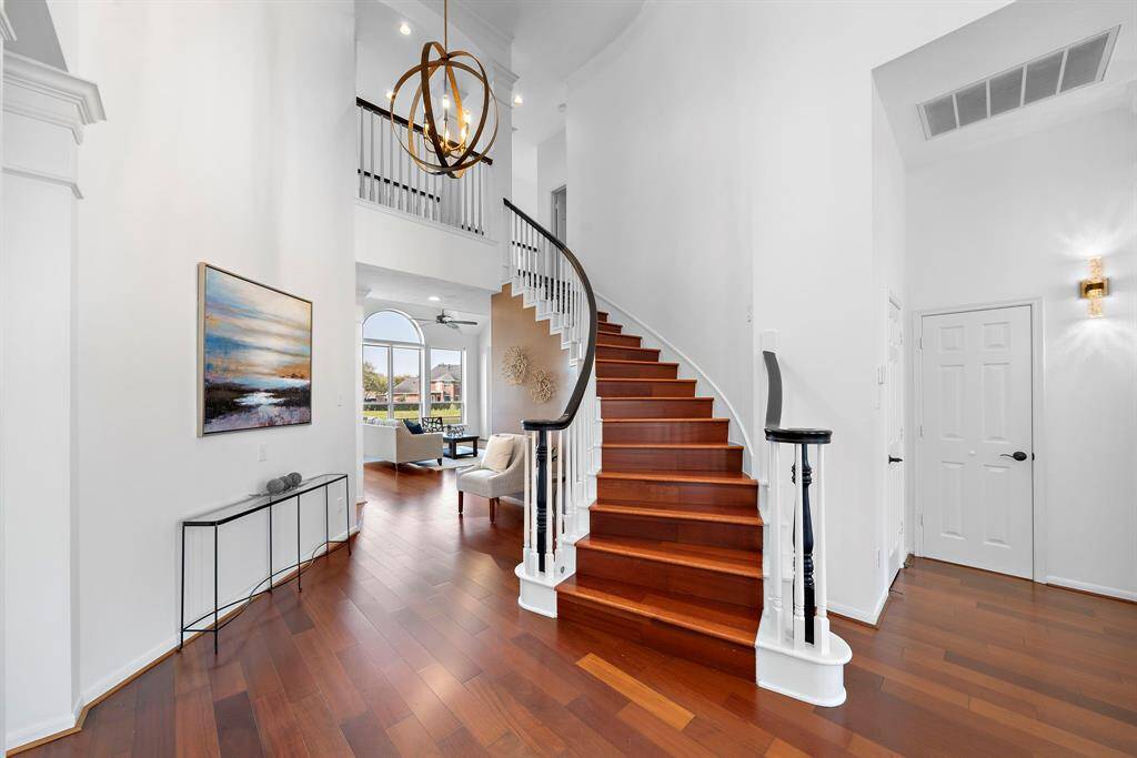 An elegant entryway with a grand curved staircase featuring rich wooden steps and white railings. The space is bright and airy, with high ceilings, modern lighting, and warm wooden flooring. A glimpse into the living area reveals large, arched windows that offer plenty of natural light.