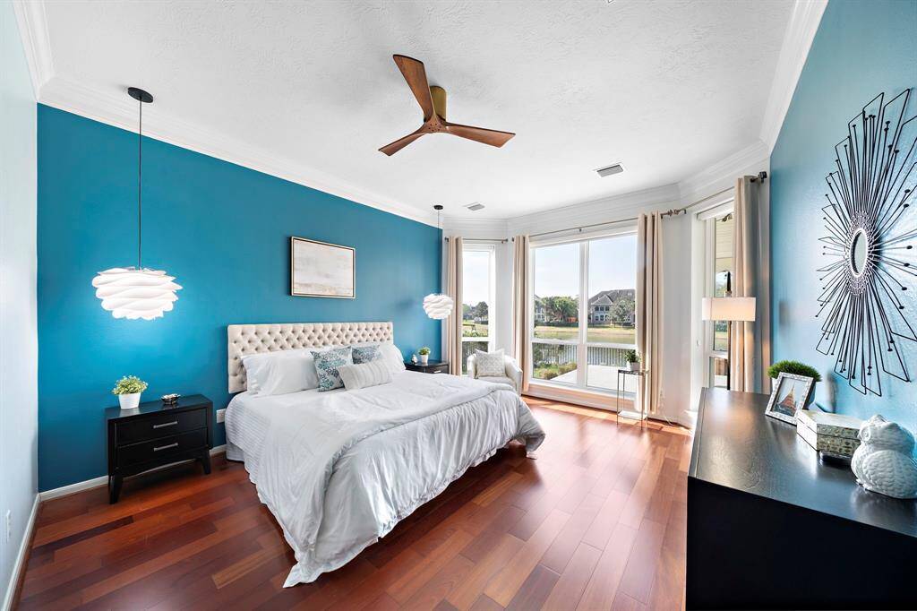This bright primary bedroom features a modern design with rich wooden flooring, a bold blue accent wall, and large windows offering a scenic water view. T