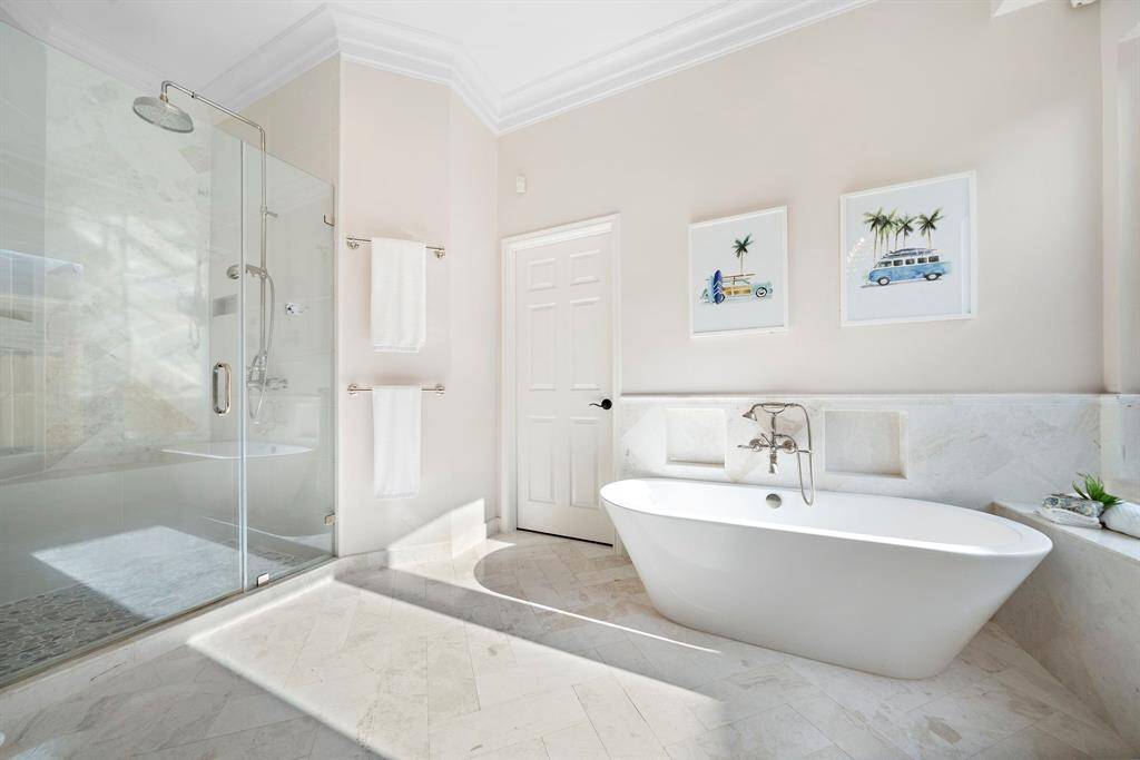 This bathroom features a spacious design with a modern freestanding bathtub and a large glass-enclosed shower. The bright space is complemented by light-colored tiles and tasteful wall art, creating a serene and inviting atmosphere.