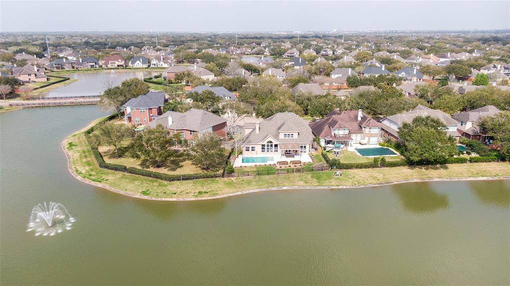 This aerial photo showcases a beautiful lakeside neighborhood with spacious homes, each featuring large backyards, some with pools. The area is lush with greenery, and a serene water fountain is visible in the lake, adding to the tranquil setting.