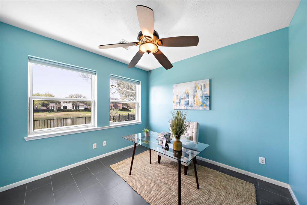 Bright home office with teal walls, large windows offering pool and lake views, and a modern ceiling fan.