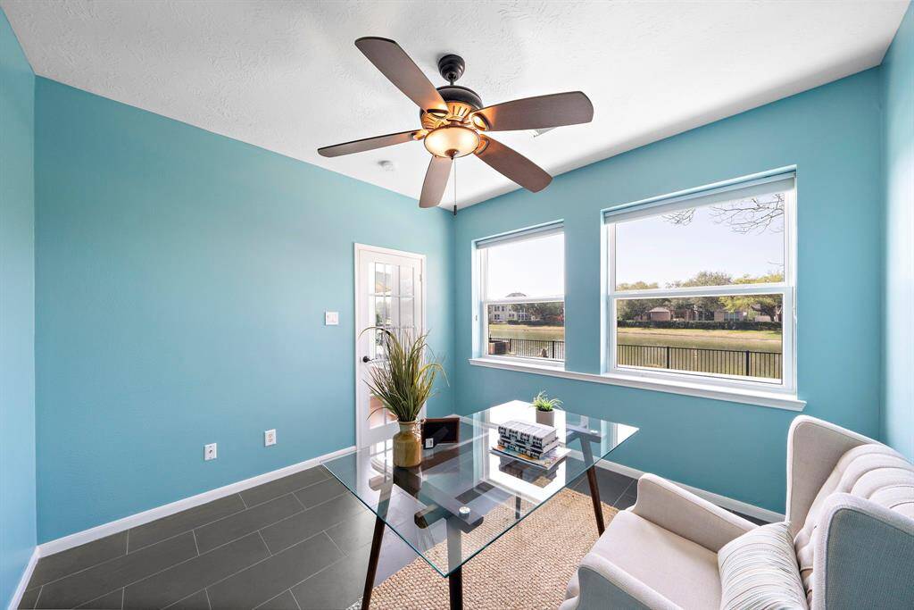 This bright and airy home office features large windows with serene water views, a ceiling fan, and a modern glass desk. Perfect for a peaceful work-from-home environment.