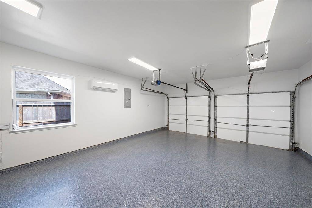 This spacious two-car garage features a clean, modern design with bright lighting, a epoxy-coated floor, and a window for natural light. It includes a wall-mounted air conditioning unit and ample storage potential.