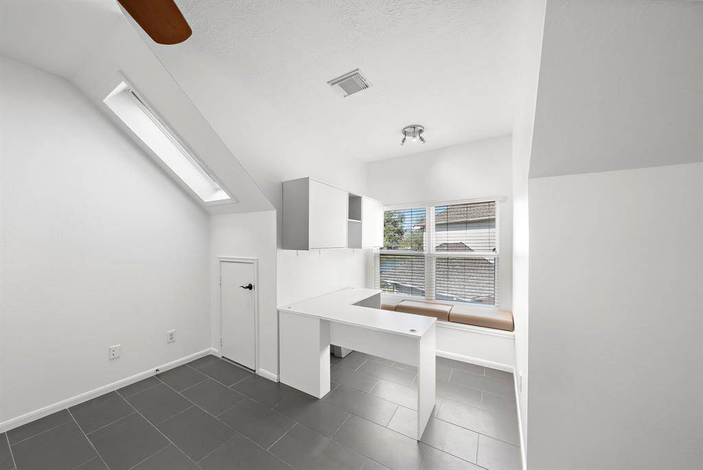 This attic room features a bright, airy space with a skylight and window for natural light, sloped ceilings, and modern gray tile flooring. It includes built-in storage, a ceiling fan, and small doors for additional storage access.