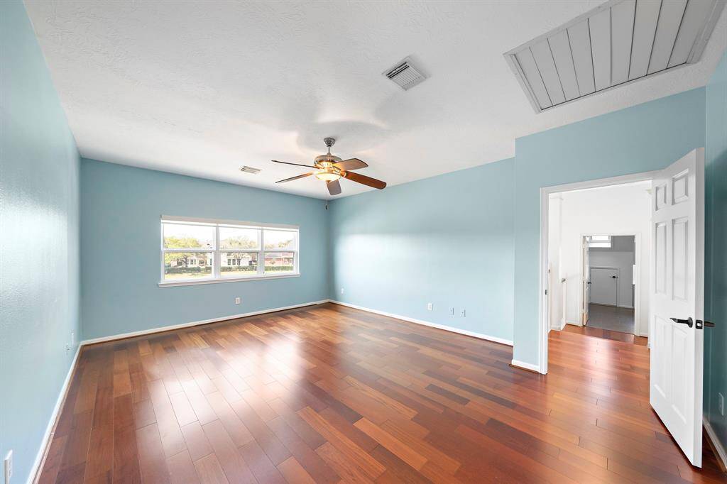 This spacious game room features hardwood floors, light blue walls, and a ceiling fan. It has ample natural light from a large window and an open doorway leading to an adjoining room.