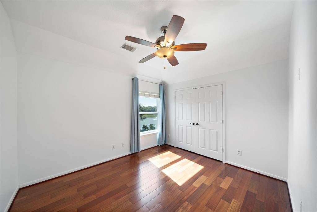 Bright bedroom with hardwood floors, a ceiling fan, and ample natural light from a large window. It features a double-door closet and neutral walls, creating a clean, airy atmosphere.