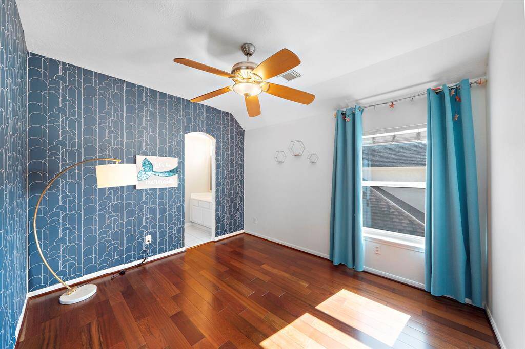 Bright room with hardwood floors, featuring a blue accent wall with a geometric pattern. It has a ceiling fan and large window with blue curtains. An arched doorway leads to another area, adding charm and functionality.