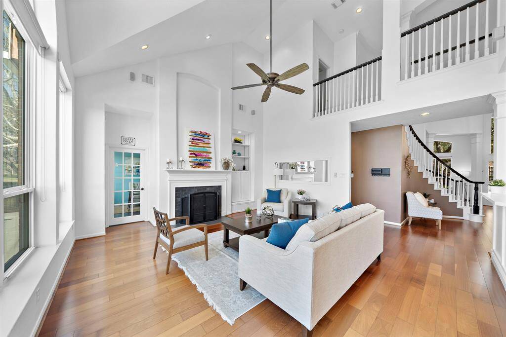 This spacious living room features high ceilings, abundant natural light from large windows. It includes a cozy fireplace, a modern ceiling fan, and a stylish staircase leading to an upper level, creating a welcoming and airy atmosphere.