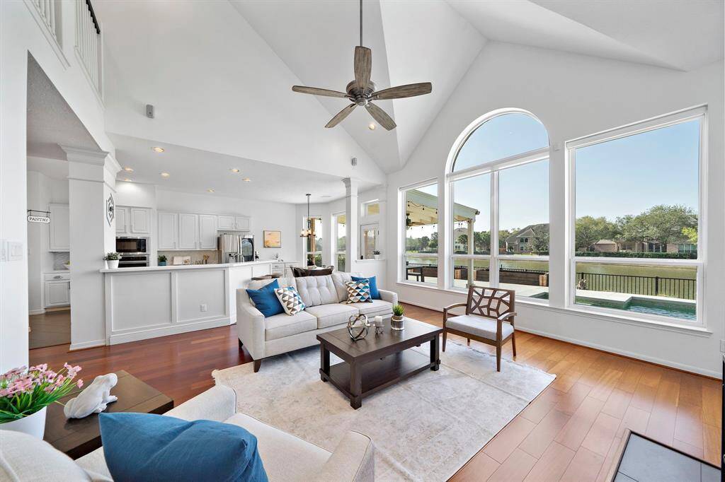 This bright, open-concept living space features high ceilings, large windows with lake views, and a modern kitchen. Wood floors and a neutral color palette create a warm, inviting atmosphere perfect for relaxing or entertaining.