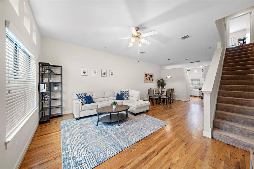 An expansive living space showcases an open layout with hardwood floors, seamlessly flowing into a dining area and kitchen. Natural light fills the room, highlighting the neutral palette and versatile design ideal for entertaining.