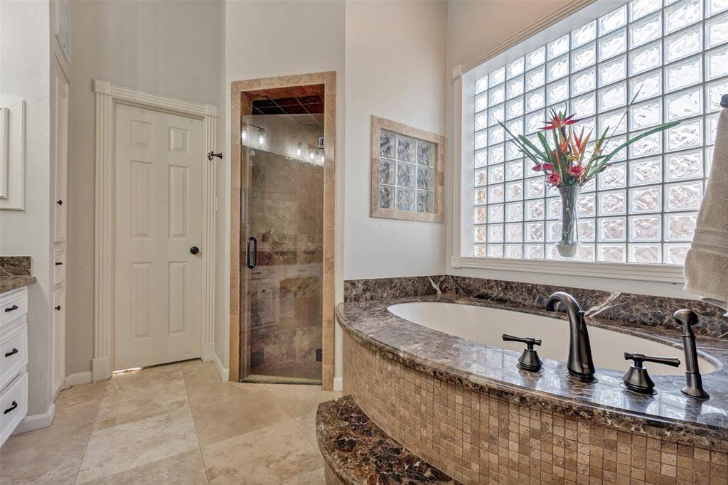 The owners' ensuite bathroom features a luxurious soaking tub with a marble surround, a glass-enclosed shower, and elegant tile flooring. A large glass block window provides ample natural light, complemented by a modern faucet and cabinetry for a sophisticated touch.