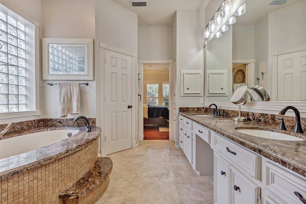 The spacious ensuite bathroom featuring a luxurious marble-topped vanity with dual sinks, a large soaking tub with a matching marble surround, abundant cabinetry, medicine cabinet, linen closet and elegant lighting. The room is brightened by glass block windows.