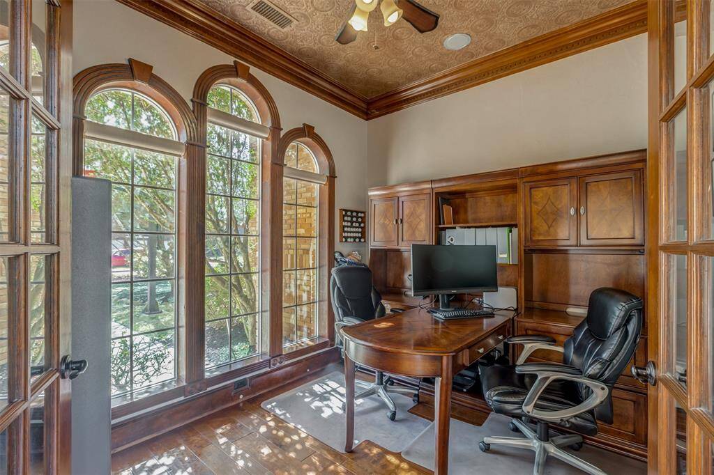 The home office boasts large arched windows, letting in plenty of natural light. It features elegant wood French doors, moldings and trim, built-in cabinetry, complemented by a decorative ceiling and hardwood floors, creating a sophisticated workspace.