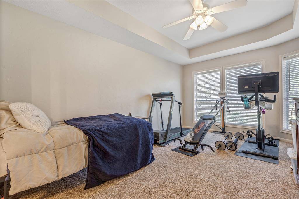 This spacious and bright bedroom features carpet, a ceiling fan, and large windows letting in natural light. It's currently set up as a home gym with a treadmill, exercise bike, and weights, alongside a cozy bed, ideal for versatile use as a guest room with an ensuite bath.