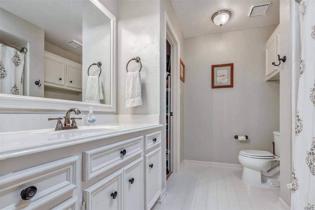 This bathroom features a clean and bright design with white cabinetry and walls. It includes a large mirror above the sink, a toilet, and shower with a patterned curtain. The space is well-lit with overhead lighting and offers practical storage solutions.