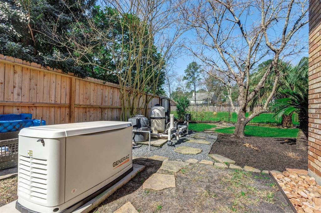 This backyard features a backup generator, with side yard wooden privacy fence and wrought iron fence in back. The mix of greenery, including trees and neatly landscaped areas, offers a serene outdoor setting.