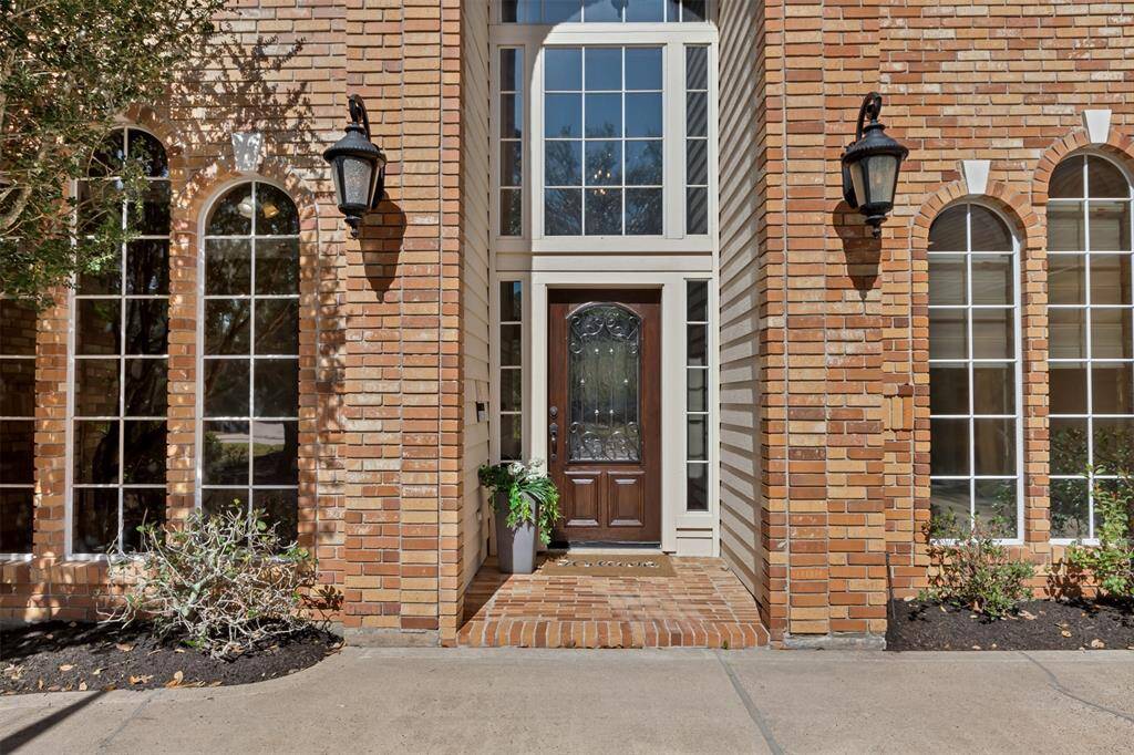 The elegant entrance features a brick facade and tall, arched windows. The front door is accented with decorative glass, flanked by sidelights, and illuminated by classic black lanterns.