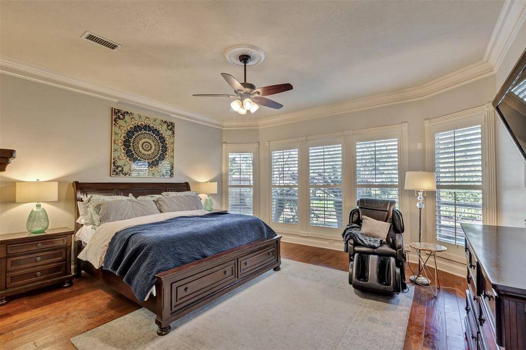 The spacious primary bedroom/owner's suite has abundant natural light through multiple windows with plantation shutters, and a cozy sitting area. Crown molding and the fan with ceiling medallion add to the room's comfort and style.