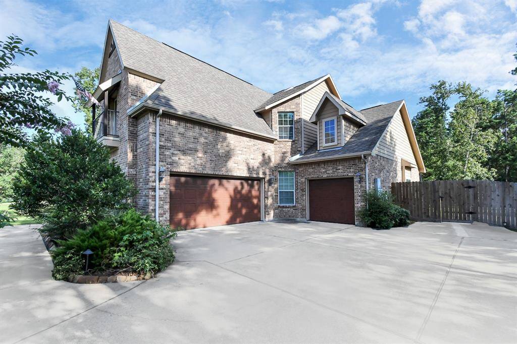This is a two-story brick home with a steeply pitched roof, featuring a two-car garage, mature landscaping, and a fenced backyard, set in a serene, green environment.