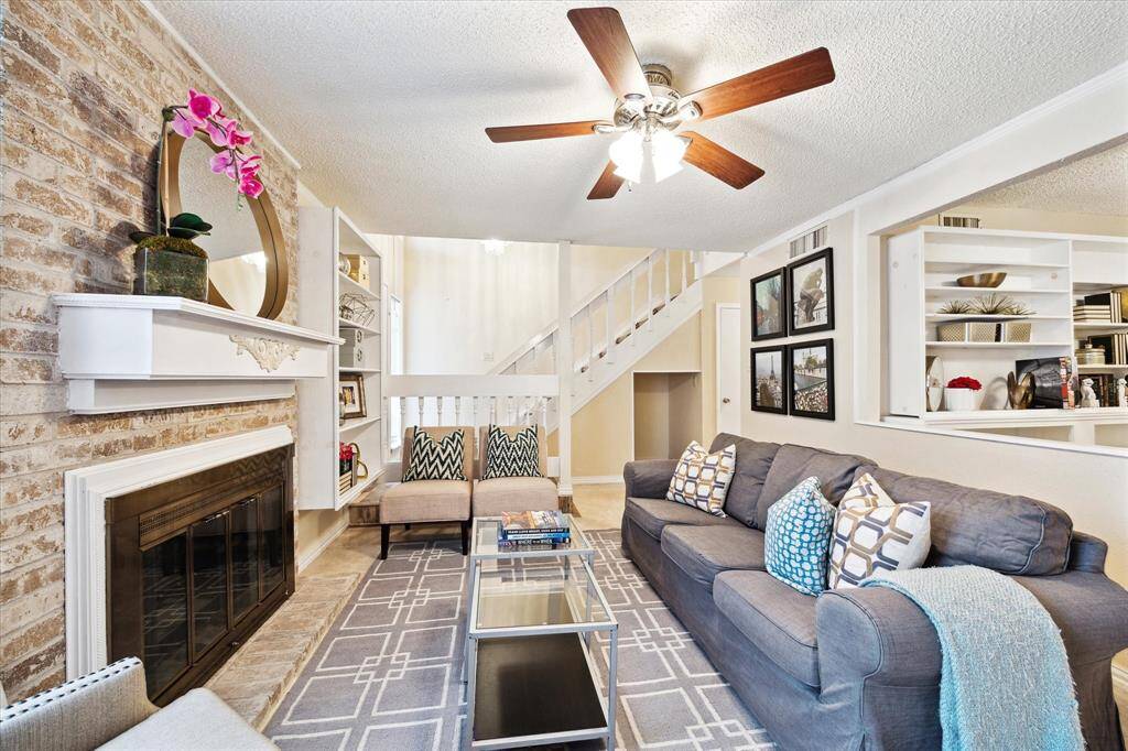 This is a bright, inviting living room featuring a large window for natural light, a charming brick fireplace, tile flooring, built-in shelves, and a staircase leading to the second level, offering a blend of coziness and elegance.