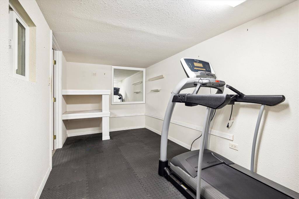 This flex space or exercise room features a treadmill and built-in shelving, with ample space. The room has a neutral color scheme and rubber flooring for durability and traction.