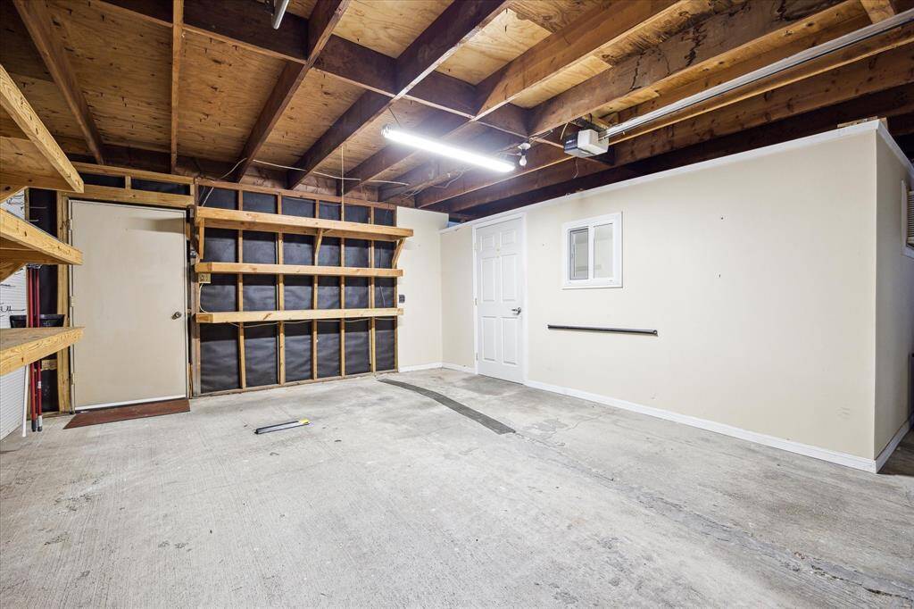 This is a spacious, unfinished garage with ample storage options, featuring exposed wooden beams and built-in shelving. It includes a side door and is well-lit, offering potential for various uses or further customization.  The structure on the right is currently used as a flex space/exercise room.