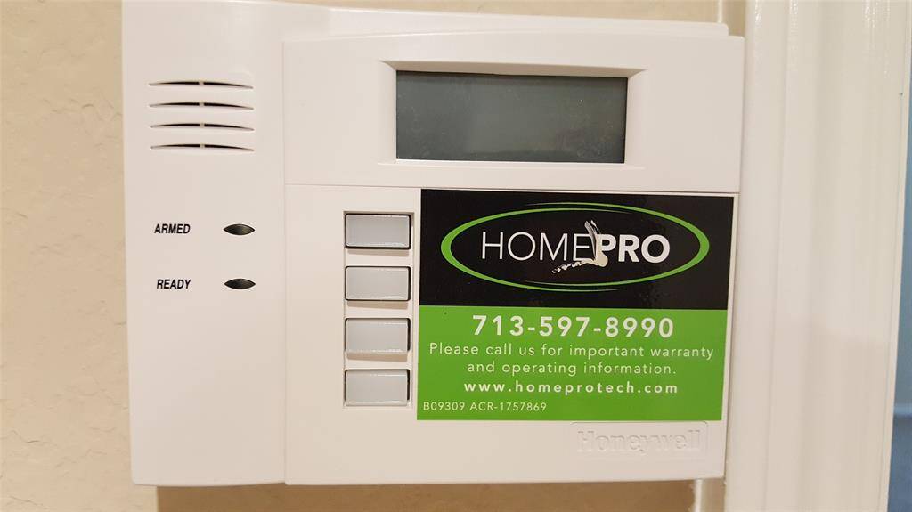 This photo shows a Honeywell security system keypad installed on a wall, featuring operational buttons and a display screen. It includes contact information for HomePro, indicating available support for warranty and operation inquiries.