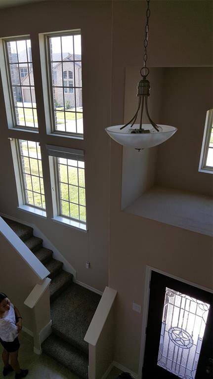 This photo shows a bright, airy entryway with a high ceiling and large windows, allowing for plenty of natural light. There's a modern hanging light fixture and a carpeted staircase leading to the upper level.