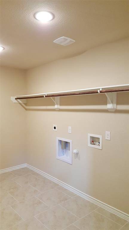 This is a spacious laundry room with neutral walls and tiled flooring. It features overhead lighting, hookups for a washer and dryer, and a sturdy wooden shelf for storage.
