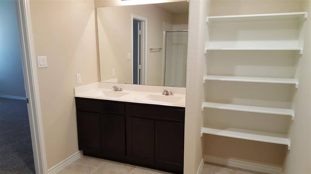 This bathroom features a double sink vanity with dark wood cabinets, a large mirror, and built-in shelving. The neutral tones create a clean and modern look, with a glimpse of a shower and bedroom in the background.