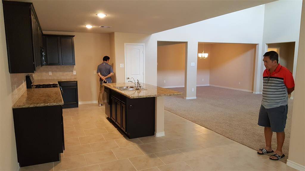This photo shows a modern open-concept kitchen and living area. The kitchen features dark cabinetry, granite countertops, and a central island with a sink. The space transitions into a carpeted living room with a hanging light fixture, offering a spacious and versatile layout.