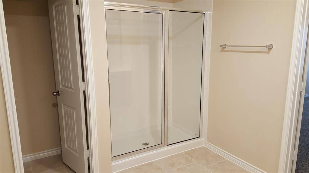 This photo shows a bathroom with a clean, glass-enclosed shower, beige walls, and tiled flooring. It features a towel rack and a door leading to an adjacent space.