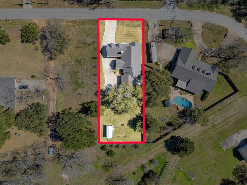 This aerial photo shows a spacious property outlined in red, featuring a large house with a driveway and ample yard space. The surrounding area is green and residential, with neighboring homes nearby.