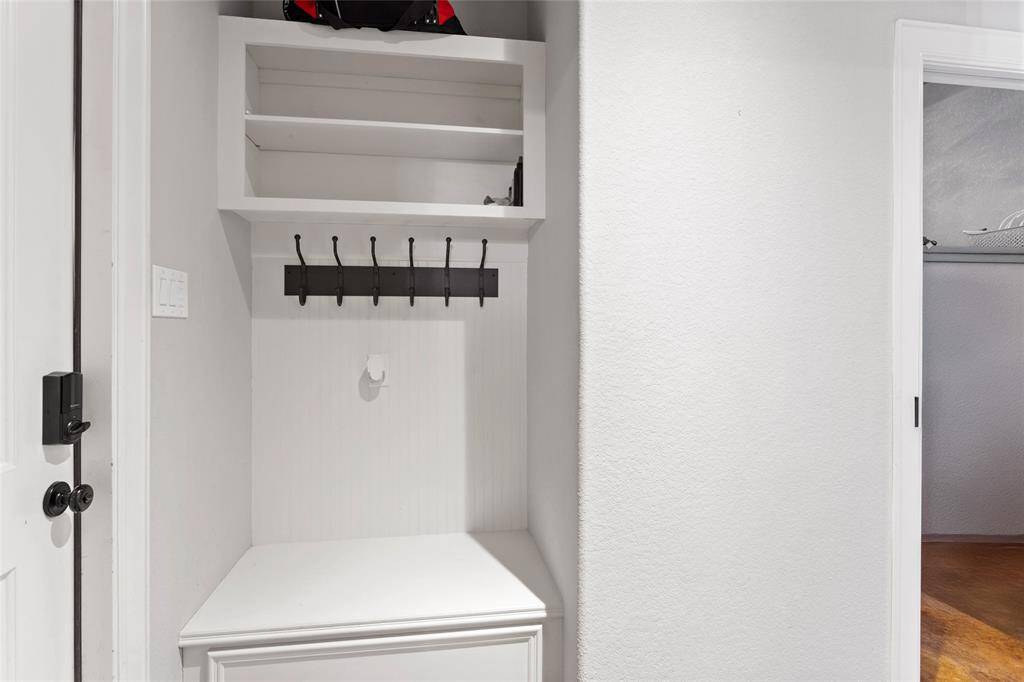 This tidy entryway drop-zone is right inside the garage door with a built-in bench and shelving. It features a row of hooks for hanging items, making it a practical space for organizing coats, bags, or accessories.