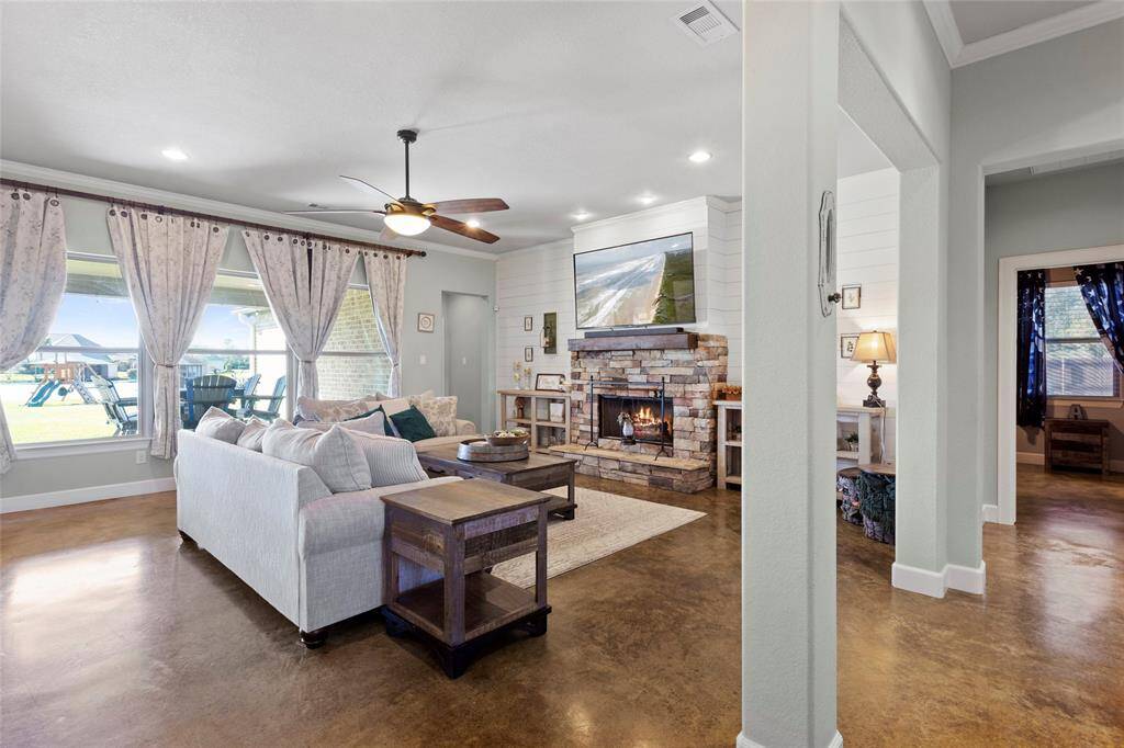 This beautiful living room features an open layout with concrete polished floors, a cozy stone fireplace, and large picture windows offering plenty of natural light and views of the lake. The shiplap accent wall adds a touch of country charm to the custom home.