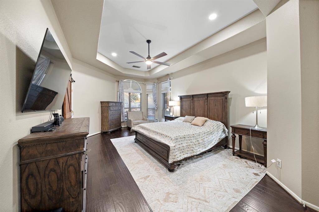 The serene primary bedroom features elegant tray ceilings, dark hardwood floors, and expansive windows that fill the space with natural light, creating a peaceful retreat for relaxation.